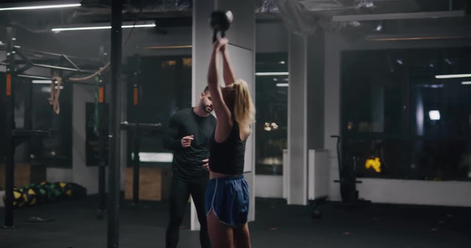 A woman repeatedly lifts a kettlebell over her head as a personal trainer watches and instructs her in a gym at night