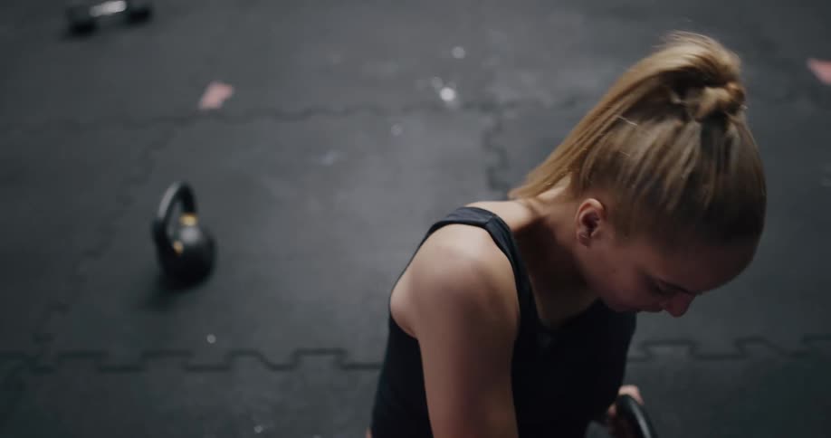 A woman exercises intensely with a kettlebell while sitting on the floor of a gym before stopping to catch her breath