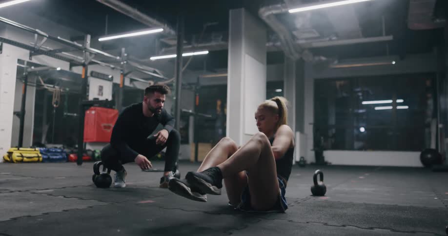 A woman exercises with a kettlebell on the floor of a gym at night as a man squatting nearby watches