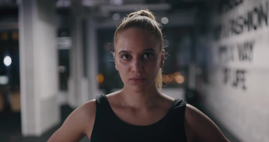 A woman in a fitness gym at night raises her gaze to stare into the camera with a serious expression