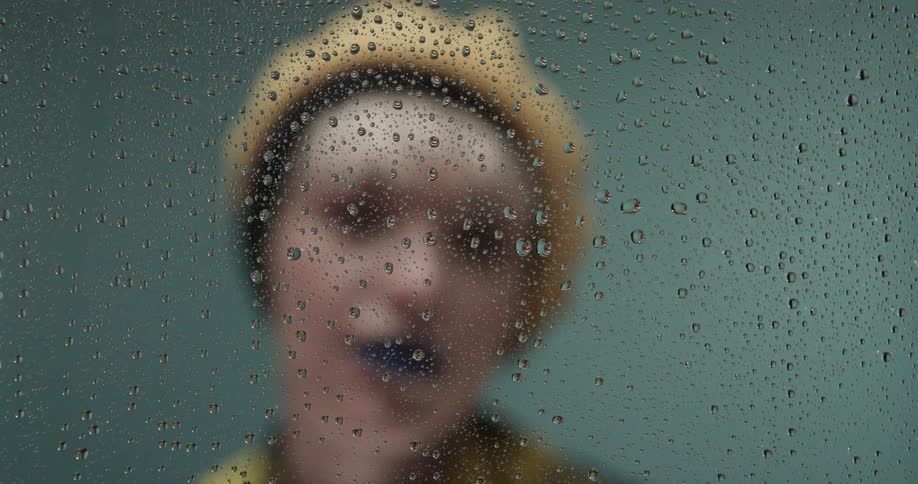 A woman with blue lipstick and wearing a yellow hat and shirt approaches a glass pane that has water droplets