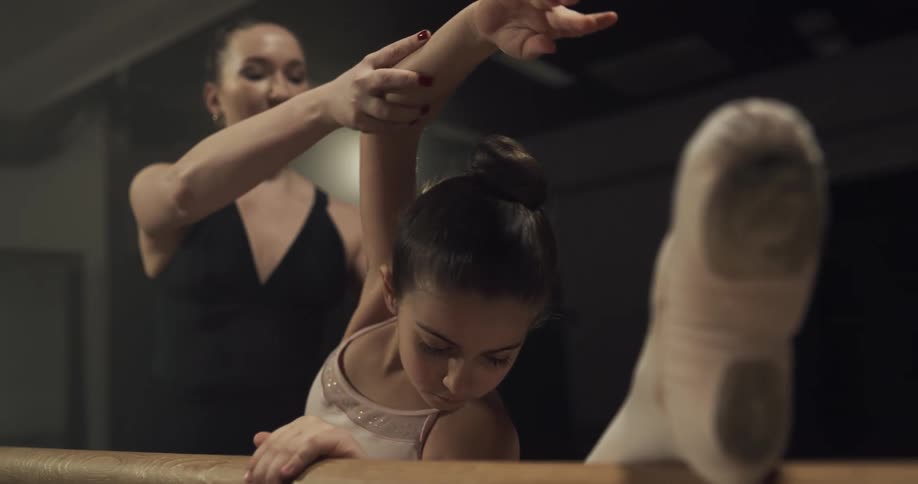 A young female ballet student is in a pose at a barre with a woman instructing her