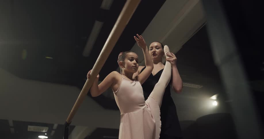 A ballet instructor helps a young ballerina to take poses against a barre in a ballet studio