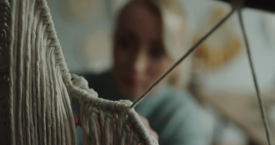 A woman wearing a blue sweater uses her hands to tie white rope in macrame