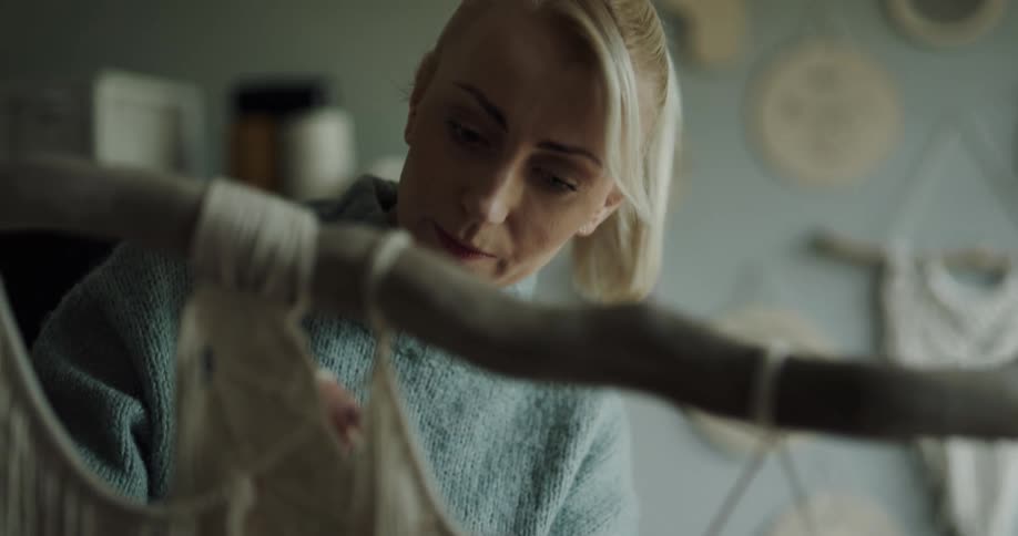 A woman weaves thick strings into an artwork hanging from a wooden branch indoors during the day