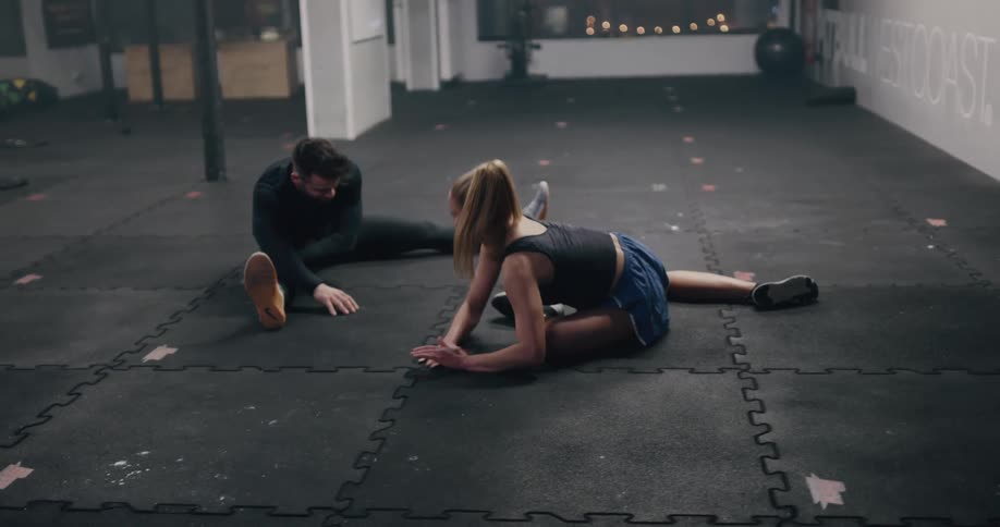 A man speaks to a woman as they do stretches on the floor of a gym while facing each other at night
