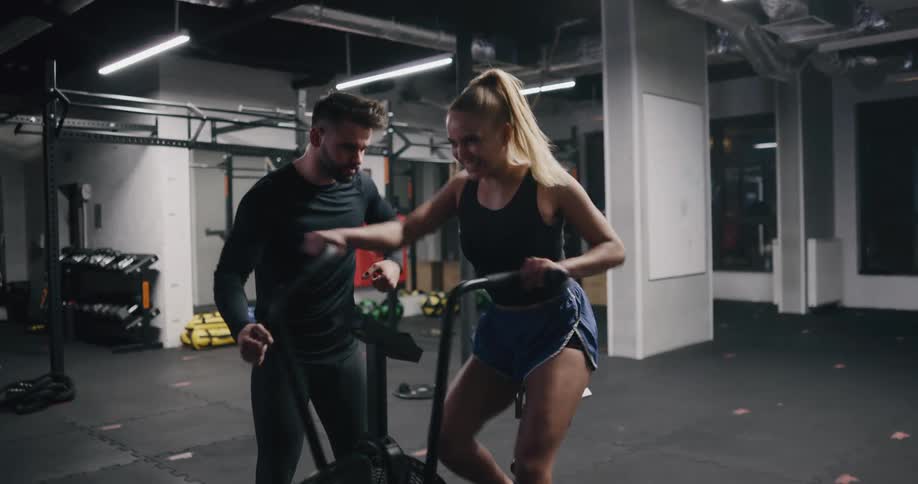 A woman gets on an exercise bike at a gym and is pushed by her personal trainer to go faster
