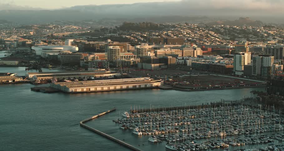 A body of water with a marina with boats with buildings a mountain range the sky and clouds in the background