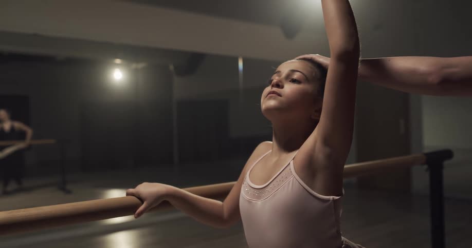 A woman instructing a girl to balance on a ballet barre in a dance studio with mirrors and a door in the background