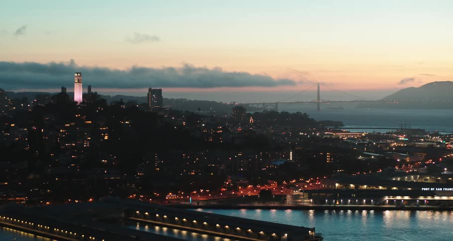 Building lights contrast with darkness of city bordered by water with fog covered suspension bridge in background under red sky
