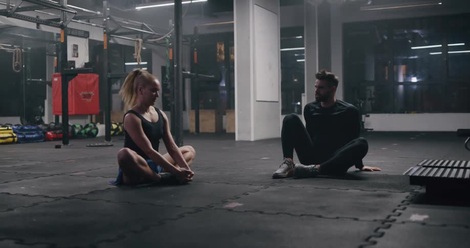 A man and a woman sitting in a room stretching with exercise equipment in the background