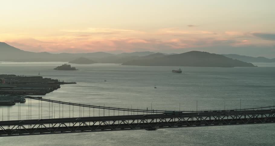 Traffic moves across a bridge over the water with hills on the horizon at sunset