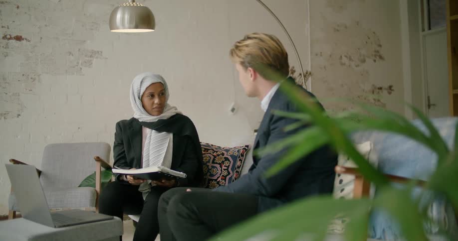 A woman holding a notebook speaks with an older man sitting beside her on a sofa in the living room of a house