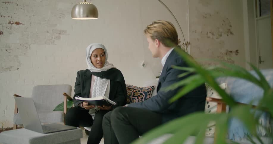 A woman holds a notebook as she talks to a man during a meeting in a casual setting with a laptop on the table between them