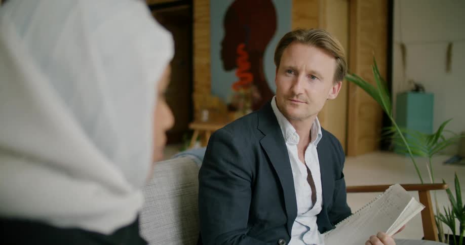 A man holding a notebook sits on a couch and talks with a woman