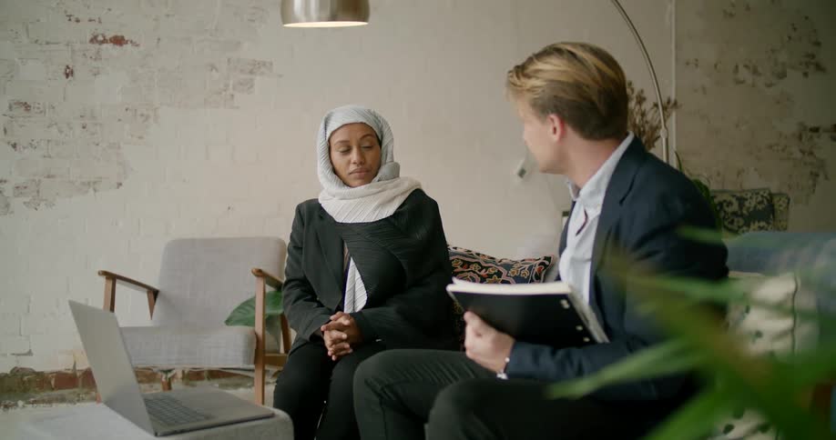 A man speaks with a client as they casually sit on a couch in front of a laptop