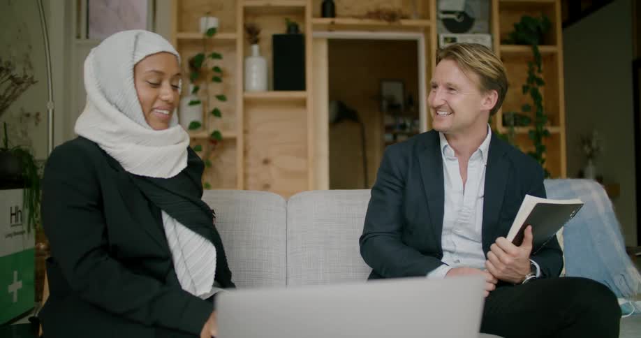 Woman wearing head covering and man holding notebook sit on couch conversing