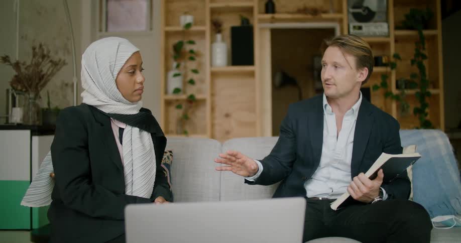 A man and woman sit on a couch and talk as the man also points at a computer screen