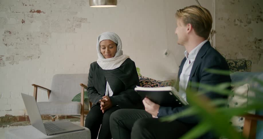 A man holding a notebook sits on a couch in front of a computer and talks with a woman