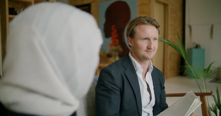 Man wearing suit sits on sofa conversing with woman wearing head covering