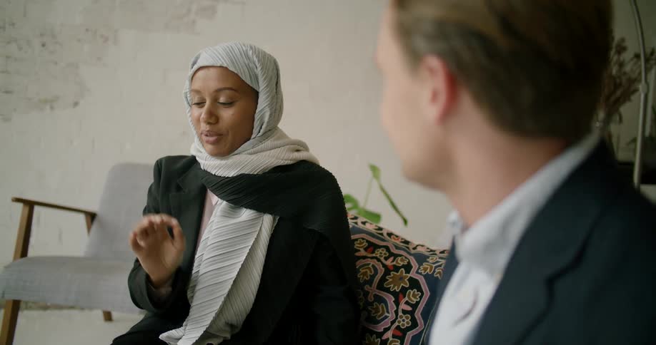 Man wearing business suit and woman wearing head covering smile as they sit and talk