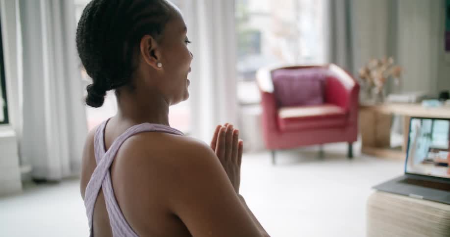 A young woman kneeling on a yoga may bows her head to greet another person practicing yoga through video chat on a camera