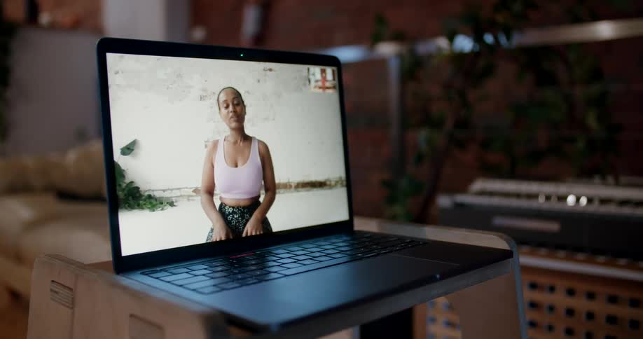An open laptop on a shelf displays a woman talking on screen in yoga attire