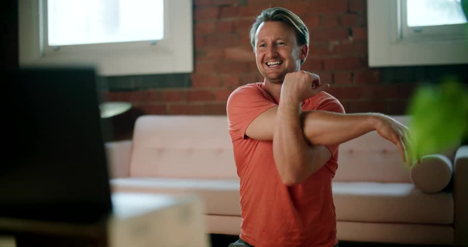 A man in a room stretching with a couch and windows in the background