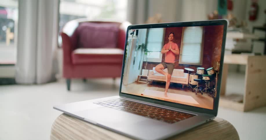 A laptop with a man doing yoga on the screen sits on a stool in a home