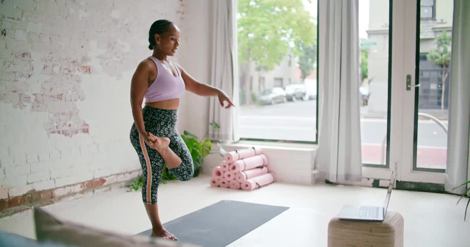 A woman speaks and gestures into the camera of a laptop as she holds a yoga pose on a mat in a room during the day