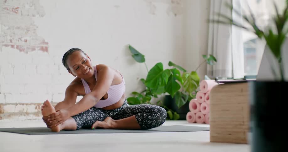 A woman in a room sitting on a mat stretching talking to a laptop