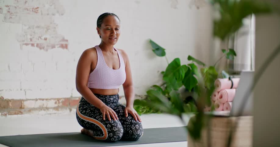 A woman kneels on a yoga mat while talking in front of an open laptop