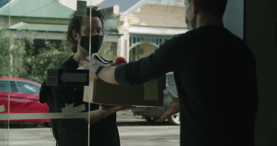 A man holding a box of produce hands it to another man who is standing indoors behind an glass door