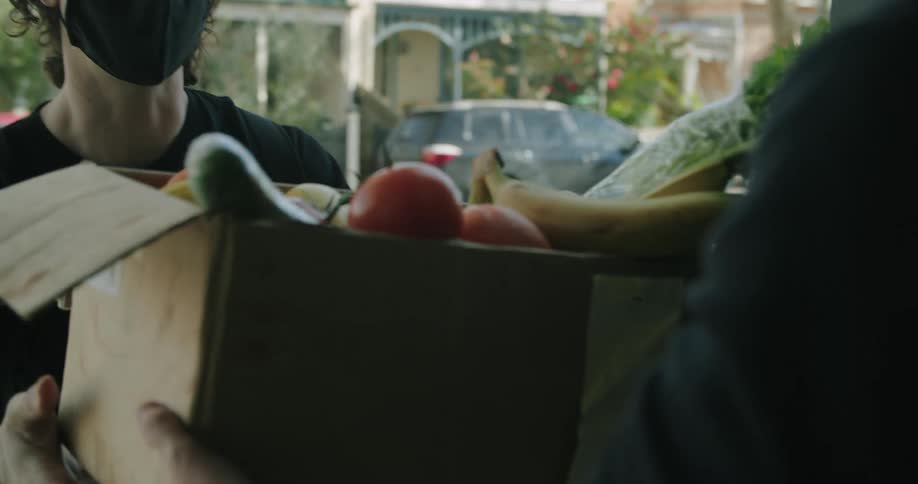 A young man in a medical mask hands a cardboard box of fruits and vegetables to another person standing in the doorway of a house before turning and walking away