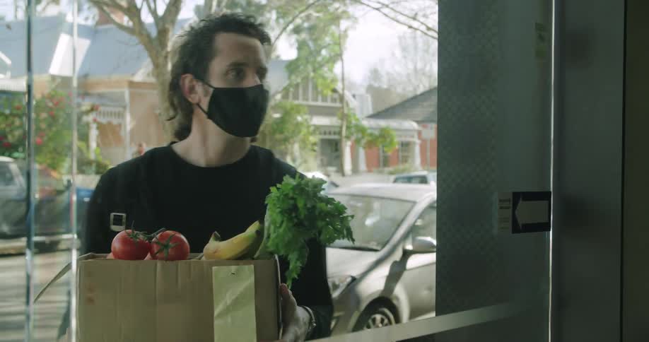 A man holding a cardboard box filled with fruits and vegetables waits for a moment outside a sliding glass door before the door opens and he hands the box off to a man on the other side