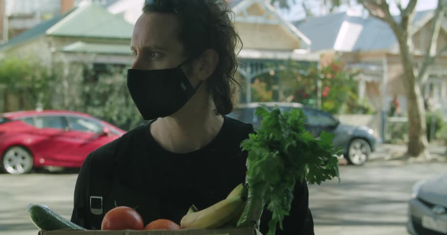 A man delivering groceries stands on a neighborhood street with a box of produce