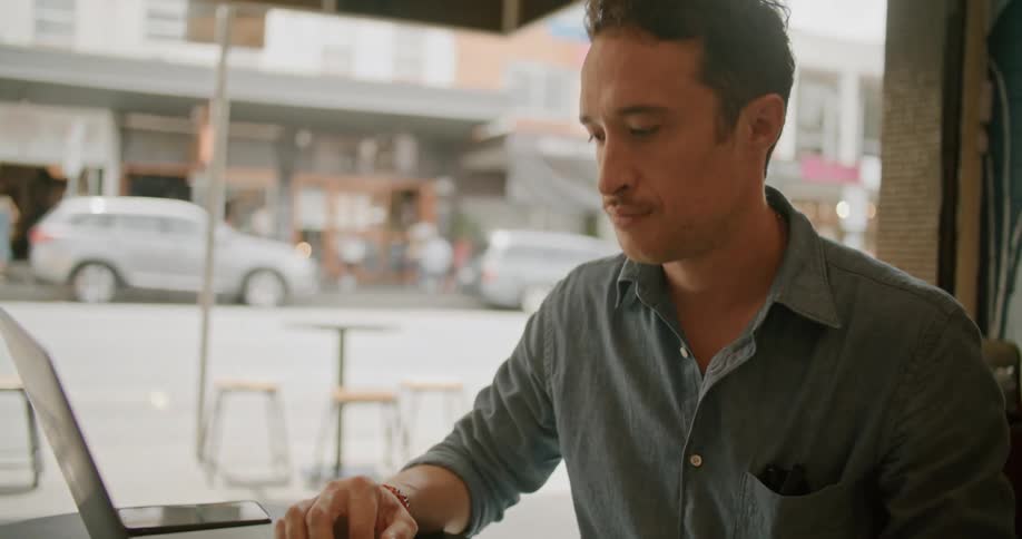 A man types on a laptop while sitting at a small table in front of a window in a building during the day