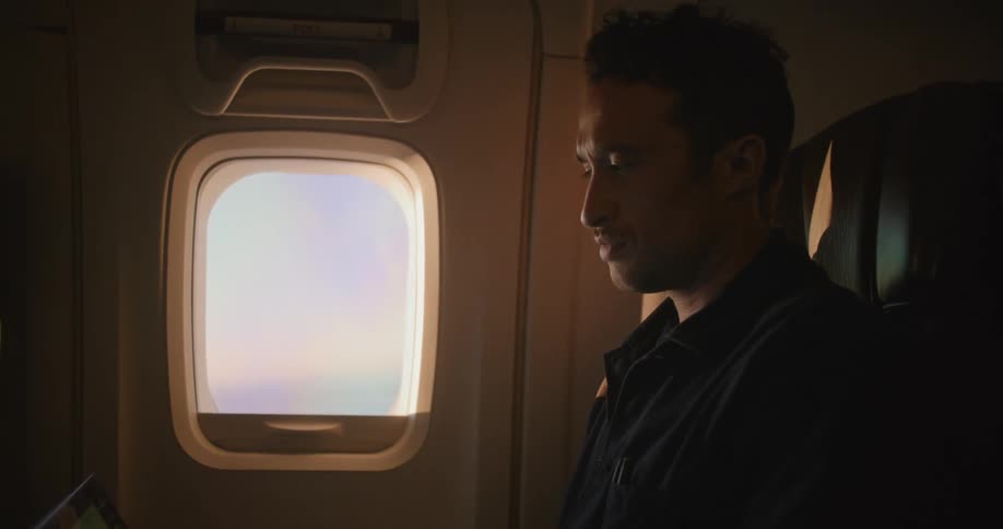 A man sitting on an airplane types on a laptop and looks out a window