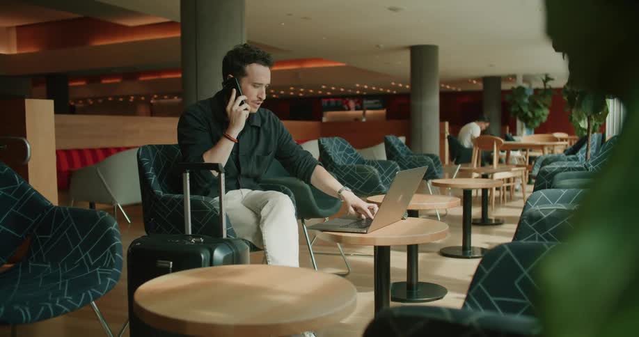 A man sitting in a chair talking on a smart phone and touching a laptop on a table
