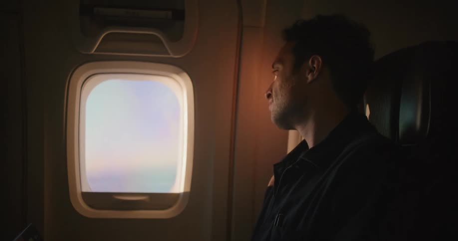 A man typing on a laptop while sitting on a plane smiles as he glances out of the window