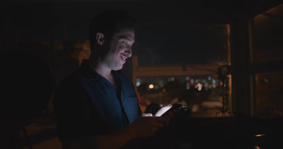 A man laughs while using his cellphone in a dark room in a building in a city at night