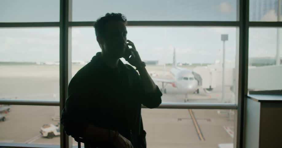 A man leaning against his luggage in front of a window in an airport dials on his smartphone before raising the phone to his ear