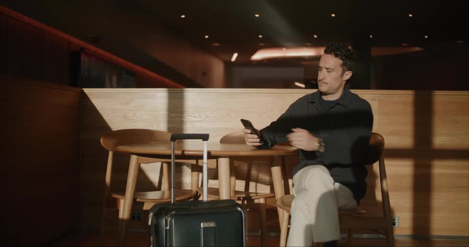 A man sitting at a table with his luggage taps his smartphone with his fingers