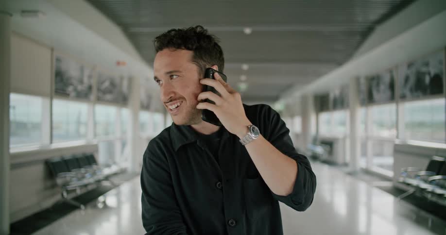 Man standing in an airport hallway with luggage answering a call on his smartphone