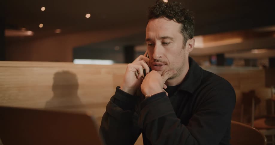 A man sits at a table with his laptop open while talking on his phone