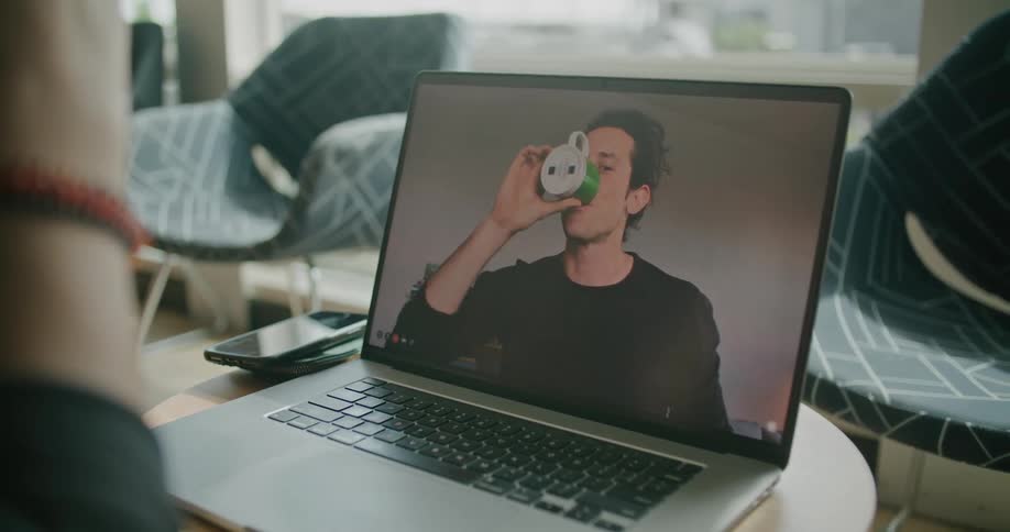 A person uses their laptop to video chat with a man who is drinking from a coffee mug during the day