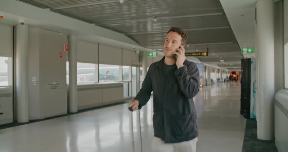 A man walking in an airport and talking on a smart phone with chairs and windows in the background
