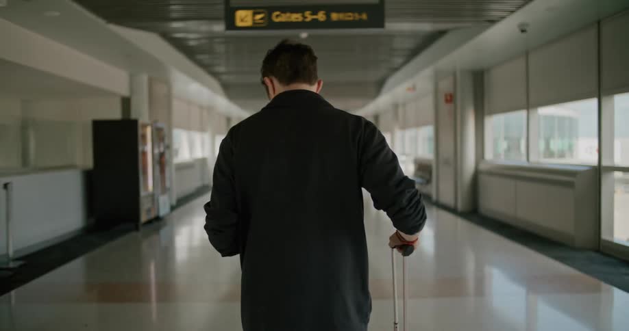 A man looks out the window then back down to his phone as he wheels his luggage in an airport