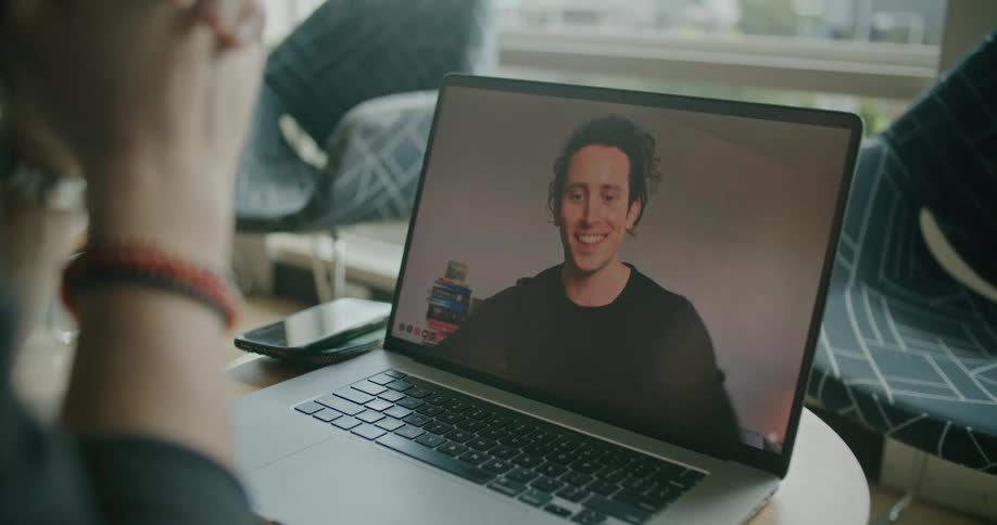 A man sits at home and looks at his laptop with another man on it and they talk