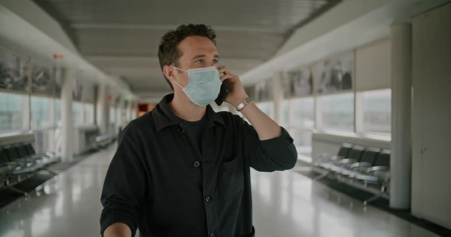 A man standing in a corridor of an airport starts a call on his smartphone before pulling his luggage along as he walks out of frame